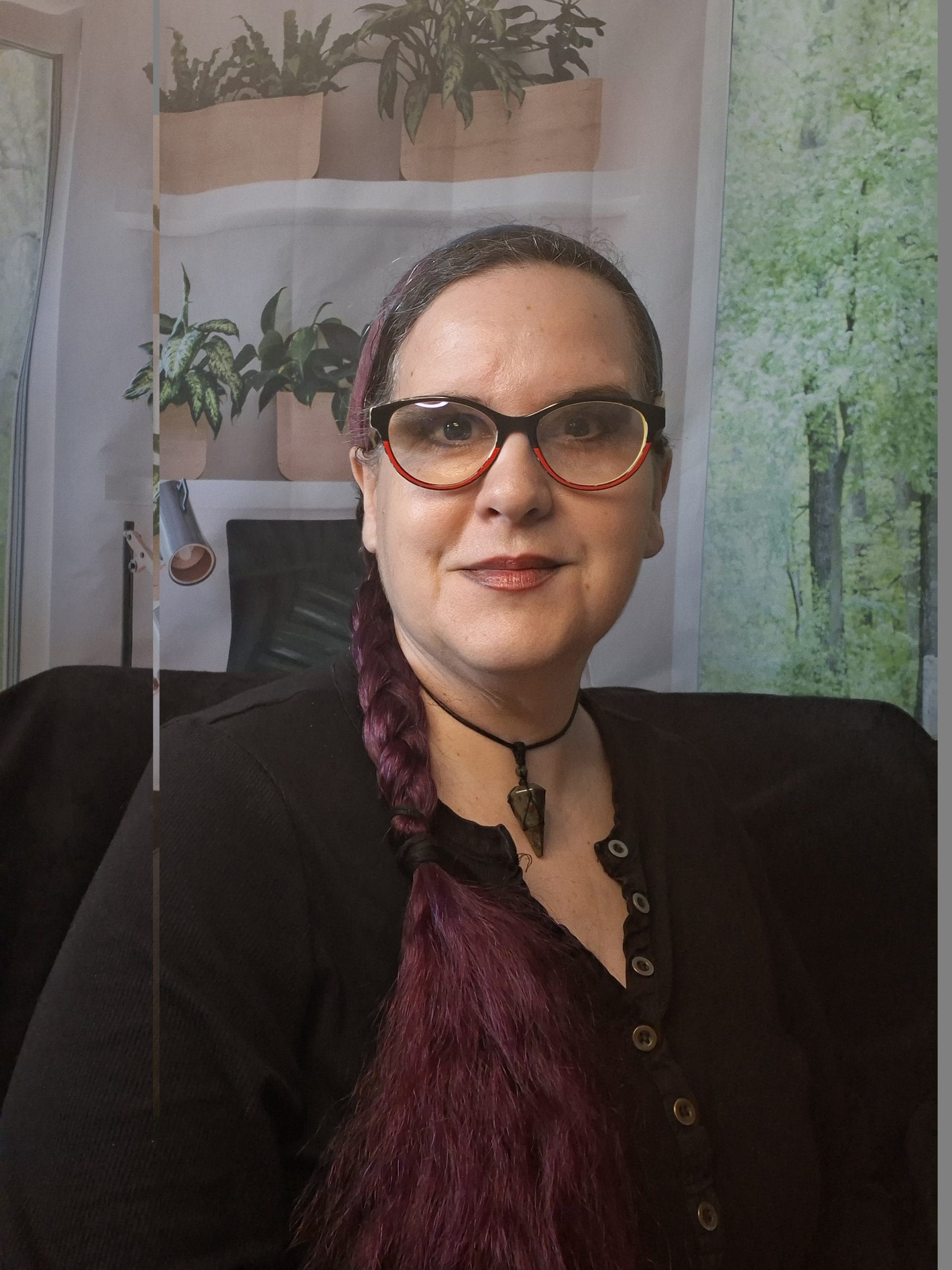TJ Mack April 2026, wearing black and read glasses looking straight ahead, sitting in front of a background showing plants and forest.
